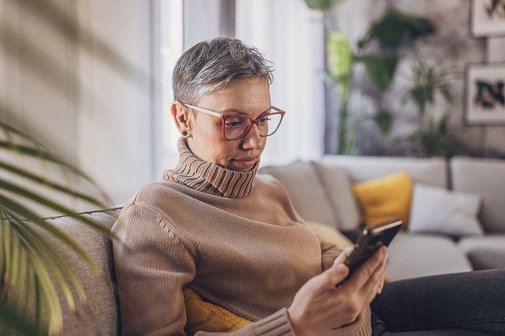 elektronische Patientenakte Erfahrung Kritik Reife Frau sitzt in ihrer Wohnung auf ihrem Sofa, sie trägt eine rote Brille und blick auf ihr Handy.