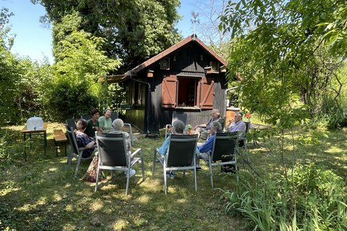 Grüner Garten mit einem Baumhaus, davor sitzen ältere Menschen in Gartenstühlen im Kreis, ins Gespräch vertieft.