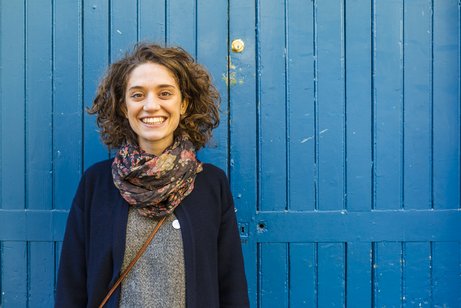 Ehrenamt beim Sozialverband VdK Baden-Württemberg Junge Frau mit lockigem Haar steht vor einem blauen Holzlattenzaun und lacht in die Kamera